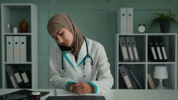 医師女性ムスリム女性医師ヒジャーブ語で携帯電話で話すことは 患者の遠隔会話への医療処方を説明します治療を書きます領収書は 健康チェックリストに記入ノートを作りますMed保険 — ストック動画