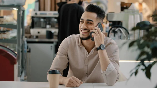 30s happy smiling laughing bearded Indian Hispanic ethnic man multiracial businessman worker entrepreneur employer sitting at cafe table talking mobile phone speak with smartphone talk with cellphone