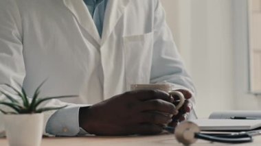 Tanınmayan beyaz önlüklü erkek doktor klinikte oturuyor Afrikalı profesyonel pratisyen doktor çay içiyor, kahve molası veriyor iş gününden sonra sıcak içecek tadıyor dinleniyor.