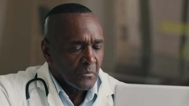 賢明な経験豊かな50歳男性アフリカ系アメリカ人医師一般開業医病院キャビネット研究室に座るノートパソコン検索医療ウェブサイトを使用して仮想遠隔医療 — ストック動画