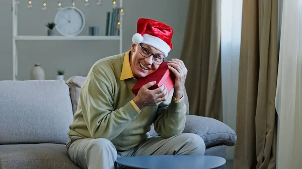 Old Caucasian senior mature grandpa 60s man grandfather male in Christmas X-mas Santa festive red hat at home holding hugging embrace present parcel gift box for New Year celebration looking at camera