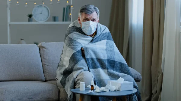 自宅でソファに座っている医療用マスクの病気の老人毛布に包まれた白人の高齢者は カメラのCovid 19流行コロナウイルスの流行インフルエンザを見て 不健康な病気成熟した高齢者を感じます — ストック写真