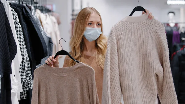 Young girl in medical mask choose clothes in store woman consumer make choice between two sweaters collects new fashionable image choosing jumpers thinking doubts lady shopping during pandemic covid19