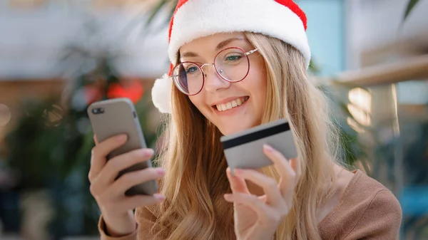 サンタの帽子の白人の女の子は 電話やクレジットカードを使用してインターネット注文を行います千年紀の女性は喜んで新年の贈り物を購入笑顔オンライン使用アプリモバイル銀行若い女性はクリスマスイブの販売を楽しむ — ストック写真