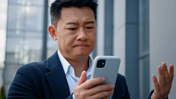 Sad Asian middle-aged adult man mad unhappy businessman feeling annoyed with using broken smart phone low battery problem angry having problem with mobile telephone frustrated bad news outdoors. High