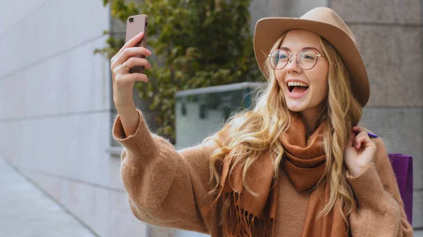 Laughing smiling female shopper buyer client girl talking video call make photo with mobile phone Caucasian woman on street with shopping bags lady with hat and glasses outdoors talk conference chat