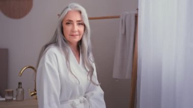 Güzel bir portre. 60 'ların beyaz, 50' lerin yaşlı, olgun kadını tuvalette duruyor. Gri saçlı büyükanne kadın bornoz giyip otel banyosunda poz veriyor nemlendirici cilt bakımı yapıyor.