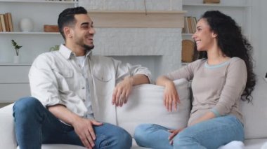 Married multiracial multiethnic interracial couple Indian Hispanic man husband and Caucasian Latina wife woman talking at home on sofa discussing talk agree gesture high five hands consent unity idea