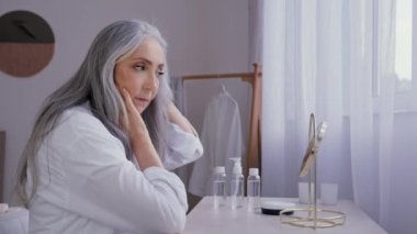 Ayna yansımasına bakan beyaz saçlı yaşlı kadın estetik cerrahi spa sağlıklı beslenme prosedürü 50 'li yaşlarda kadın model, anti-yaş kremi kozmetik ürünleri şımartıyor.