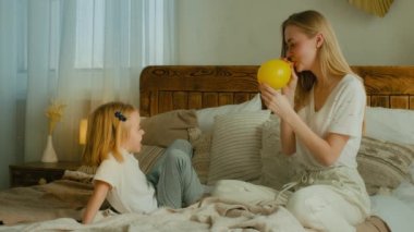 Kafkasyalı yetişkin bir anne ve kızı yatak odasında oynuyorlar anne sarı balon üflüyor şişme top içinde hava hazırlıyor doğum günü şenliği için küçük kız çocuğu annesiyle eğleniyor.