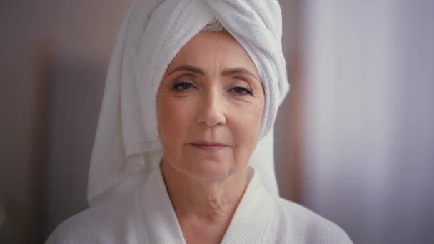 白人の高齢者の高齢者の魅力的な中年女性60代の女性50代の女性モデルは カメラを見てしわ顔で頭の上にバスローブと白いタオルを身に着けています 広告スキンケアスパ手順 — ストック動画