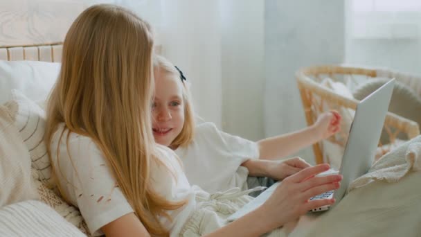 ベッドで寝そべっているラップトップのオンラインストアを閲覧している娘を持つ白人の母親は 一緒にベッドルームでインターネットショッピングウォッチ映画を購入します お母さんは小さな赤ちゃんの女の子を教えていますコンピュータサイバー空間を使用して — ストック動画