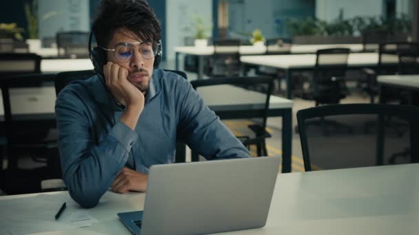 退屈疲れてアラビア系インド人の男性ビジネスマンは オフィスの机に座って疲れ怠惰な悲しい男性でヘッドフォンで退屈なオンラインウェビナーを聞く仕事の日の終わりについてのモチベーションの夢の退屈不足を感じる — ストック動画