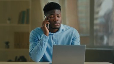 Afrikalı erkek iş adamı, etnik 30 'lu yaşlarda ofis çalışanı cep telefonuyla iş konuşmalarından uzakta çalışıyor. Kablosuz telefon görüşmeleri yapıyor, bilgisayar ekranına bakıyor, akıllı telefon kullanıyor.