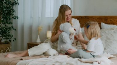 Beyaz anne bebek bakıcısı anne yatak odasında oyuncak ayıyla küçük tatlı bir kızla oynuyor. Çocuksu bebek ve anne yatakta oyuncakla oynuyorlar eğlenceli oyun oynuyorlar yeni evde sevgi bağı kuruyorlar.