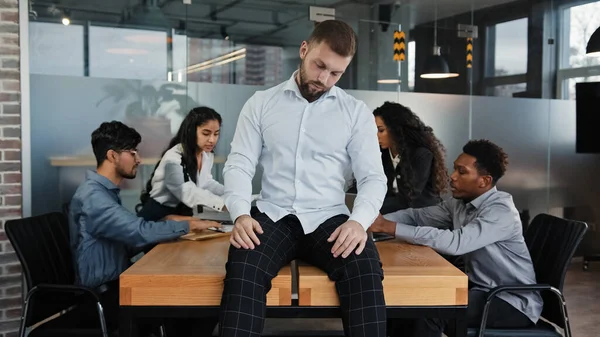 Caucasian businessman lazy man worker sitting on table with close eyes pretend sleeping tired exhausted on background of multiracial multiethnic team workers discussing project at business meeting