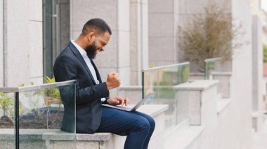 African American successful ethnic guy worker man entrepreneur executive employer ethnic businessman working with laptop outdoors near company office win victory online winning business achievement