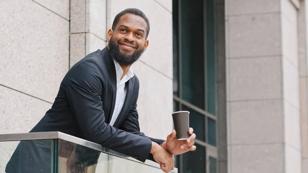 Smiling relaxed African American corporation employee company CEO office manager businessman entrepreneur ethnic man guy standing on balcony with coffee tea has work break resting dreaming outdoors