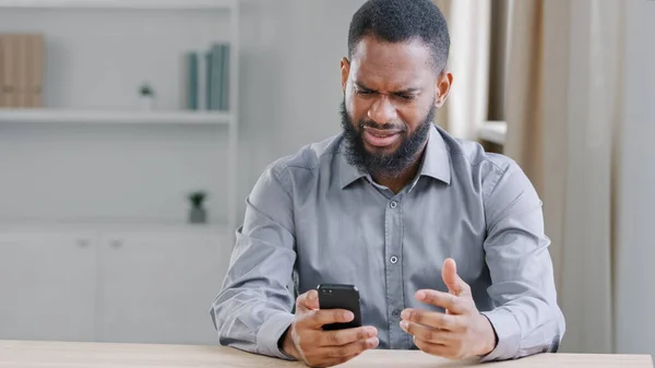 Stressed disappointed African American ethnic bearded man with mobile phone reading bad news. Office worker businessman with smartphone sad with failure online payment app error cell service problem