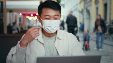 Asian man in medical mask sitting in cafe typing on laptop remote works internet happy healthy male freelancer tourist take off protective respirator smiles enjoys fresh air rejoices at end quarantine