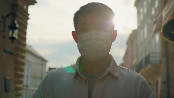 ヘッドショット若い不健康な男医療用マスクの屋外に立って太陽の下でカメラを見て責任ある男性観光客は パンデミックの健康保護コンセプト中に保護マスクを身に着けている — ストック動画