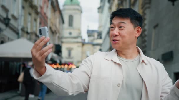 笑顔男立って屋外でビデオ通話で話す上の携帯電話幸せなアジアの男性観光ブロガーインフルエンサー記録Vlogで市内中心部で使用してスマートフォンを見てウェブカメラで携帯電話画面 — ストック動画