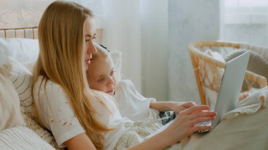 Caucasian mother with daughter browsing laptop online store lying in bed buying in internet shopping watch movie at bedroom together. Mom teach little small baby child girl using computer cyberspace clipart