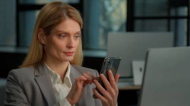 Caucasian middle-aged adult woman mature business female lady businesswoman entrepreneur scrolling browsing mobile phone win winning gesture celebrate achievement success online bet prize in office