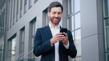 Smiling man holding phone checking email message reading good news happy businessman using mobile application on smartphone looking at camera showing thumb up gesture approval excellent result sign