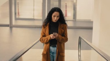 Caucasian Hispanic businesswoman with mobile phone reading e-mail in smartphone woman in shopping center on escalator. Female lady girl booking taxi car with online app in airport on moving staircase