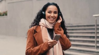 Happy carefree girl brunette woman young lady female walking in fall autumn street in city listening music earphones with mobile phone sound audio app online musical service in headphones song walk