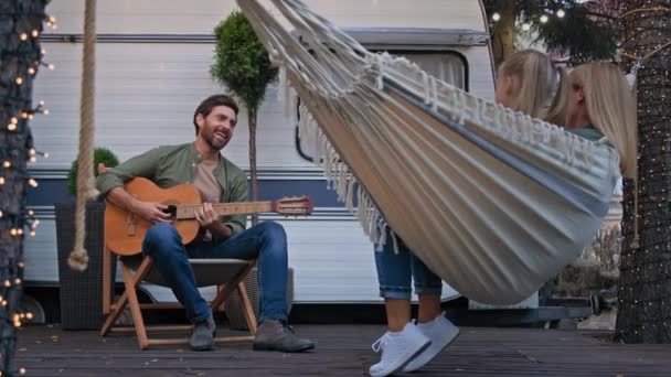 コーカシア人の家族は休暇でキャンプで休暇を過ごしていますヴァントレーラーの近くの母親と娘の女の子子供はハンモックに座って屋外で歌うお父さんがギターで面白い歌を歌うことで音楽を演奏するのを聞いて — ストック動画