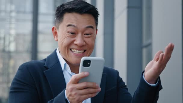 アジアの幸せな驚きの男を閉じます感情的に成功を楽しんで良いニュース40代のビジネスマンの上司は モバイルオンラインベット入札お金の賞金の勝利は屋外のスマートフォンでオファーの機会を受ける — ストック動画