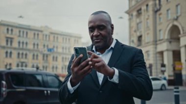 African middle-aged man entrepreneur businessman has video call mobile conference with phone in city walk street talk to smartphone chat virtual talking outdoors walking lawyer speaking web camera