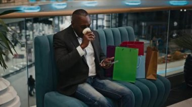 African middle-aged man male client buyer customer in shopping mall sitting on sofa with purchases typing browsing mobile phone chatting message about discount order booking goods drinking tea coffee