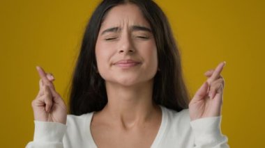 Indian girl lady in yellow studio background crossed fingers close eyes praying hope wish woman waiting miracle pray ask God for help success luck believe trust expect win beg winning dreamy wishing