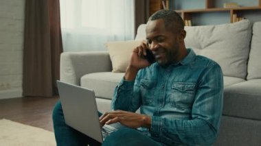 Focused busy multitasking man typing on laptop talking on mobile phone male freelancer working at home sitting in living room communicates with client using smartphone browsing information on Internet