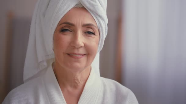 接近女性画像50多岁头戴毛巾的中年女士在浴室转过头对着相机微笑60多岁沐浴在浴衣中的成熟女人笑着对面部护肤化妆品整容满意 — 图库视频影像