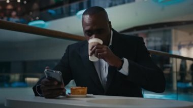 African American adult middle-aged ethnic man worker businessman employer entrepreneur eating drinking relax sitting at table in cafe drink coffee tea reading news in mobile phone smartphone browsing