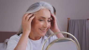 Caucasian old 60s middle-aged mature Caucasian woman 50s lady female looking at mirror reflection in morning touching gray hair enjoy wrinkled face skin pampering with hairstyle haircare moisturizing
