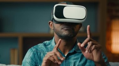 Close-up excited adult man in virtual reality glasses playing online game while sitting on couch in living room gesturing hands modern male gamer with surprised expression smiling plays in cyberspace