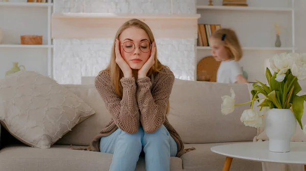 Tired upset Caucasian mother sitting on sofa at home sad frustrated stressed exhausted fatigued mom suffer headache with active noise difficult kid child baby girl daughter running around playing