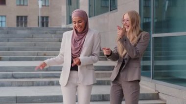 Happy smiling girlfriends diverse colleague European Caucasian blonde woman and Indian oriental girl in hijab dancing, triumphing after job interview. Successful businesswomen celebrating promotion