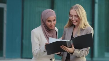 Friendly smiling young diverse female colleagues looking in document file delighted satisfied with result of the report. Indian girl in hijab and young European woman working together during break
