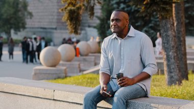 African American ethnic middle-aged man mature senior businessman male sitting outdoors in city with coffee tea and phone waiting for date business meeting looking for friend relaxing on work break