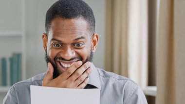 Surprised shocked ethnic winner man excited wonder male executive entrepreneur business office worker reading letter good news. Happy ethnic African American businessman read job offer win achievement