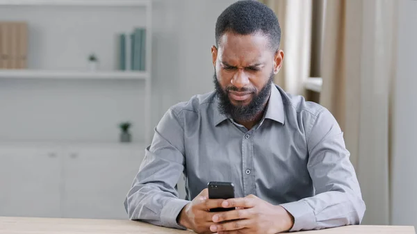 Confused puzzled angry African American ethnic bearded man boss with mobile phone reading bad news has business trouble sitting at table in office. Businessman with smartphone failure online app error