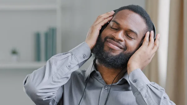 Happy relaxing calm guy at office African American man worker ethnic businessman manager entrepreneur executive in headphones listening to music at workplace with closed eyes enjoy favorite audio song