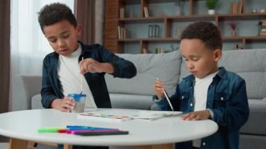 Two brothers African American children kids ethnic sons sibling painting at home together with acrylic watercolor colors little boys schoolboys indoors paint picture with paintbrushes hobby homework