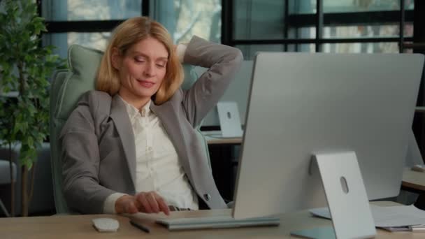 40多岁的白人中年妇女满意地完成了坐在工作场所的计算机工作 松懈的女职员下班后休息一下 双手放在脑后 — 图库视频影像